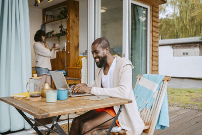 Happy mature man using laptop while sitting at table on porch outside house