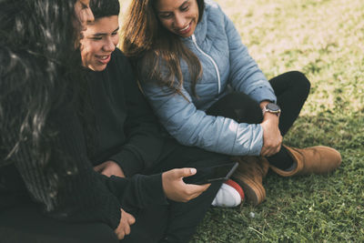 Hispanic teenager holding a smartphone sitting on the grass with his mother and sister in a park