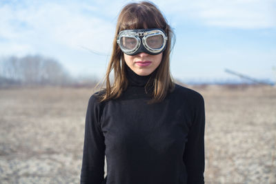 Portrait of young woman wearing swimming goggles while standing on field against sky