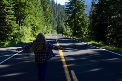 Rear view of woman with arms outstretched standing on road amidst tree