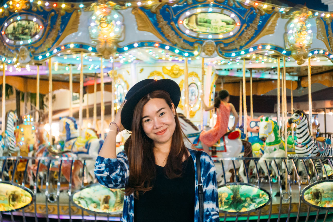 amusement park, leisure activity, one person, young adult, adult, front view, retail, carousel, arts culture and entertainment, amusement park ride, portrait, enjoyment, casual clothing, women, happiness, smiling, waist up, real people, standing, hairstyle, beautiful woman, consumerism