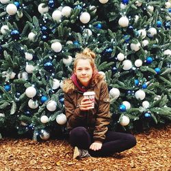 Portrait of girl holding drink while sitting on field against christmas tree