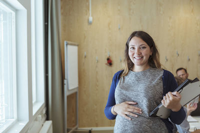 Portrait of smiling pregnant businesswoman with file and laptop standing in office