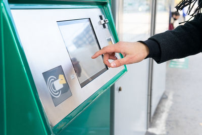 Cropped unrecognizable male in black coat standing with hand in pocket and buying ticket on railway station in city in daytime