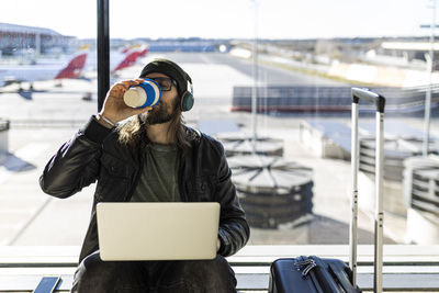 Young hipster guy in black jacket and hat with headphones drinking takeaway coffee and using laptop while sitting against glass wall in airport lounge