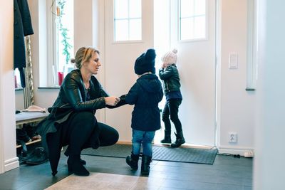 Mother helping daughter in wearing jacket while crouching at home