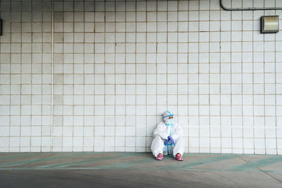Female nurse wearing protective suit with sitting against tiled wall
