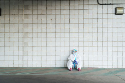 Female nurse wearing protective suit with sitting against tiled wall