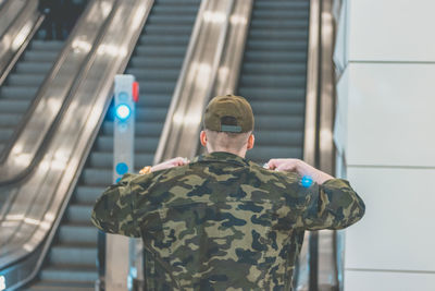 Rear view of man in camouflage jacket standing against escalator at railroad station