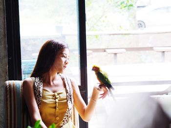 Woman looking at bird perching on her hand by window at home