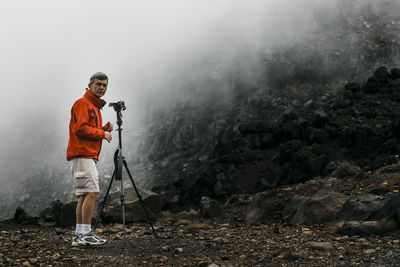 Portrait of man with camera and tripod standing against mountain during foggy weather