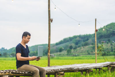 Man using laptop while sitting on boardwalk at rice paddy