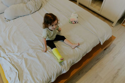 A baby girl with a cast on her leg sits on the bed.