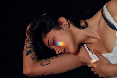 Serene female with tattoos lying on floor with closed eyes and flares on faces on black background in studio