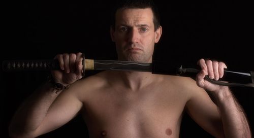 Portrait of shirtless serious man holding sword against black background