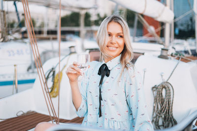 Beautiful blonde young woman in blue dress with glass of soda on boat at pier in sunset time