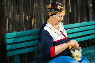 Senior woman peeling apple while sitting on bench at park