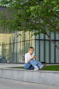 Focused young woman sitting on bench in spring park under tree outdoors resting using smartphone