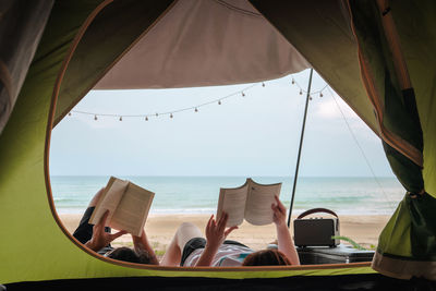 Asian couple camping and reading book on beach with sea background on summer vacation