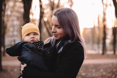 Happy young mother carrying baby son standing in park in autumn