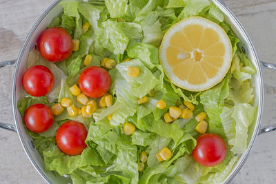 A colander with green salad, cherry tomatoes, corn and lemon. from above.