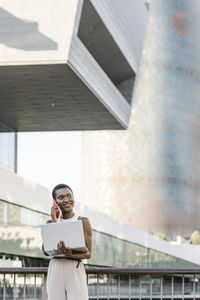 Smiling businesswoman with laptop talking on mobile phone by railing