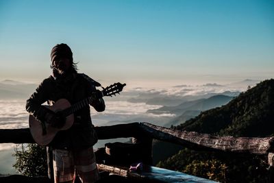 Man playing guitar on mountain against sky during sunset