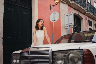 Positive female in dress standing near white old timer automobile on street with shabby building in city