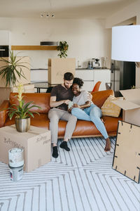 Multiracial couple sharing smart phone while sitting on sofa in living room at new home