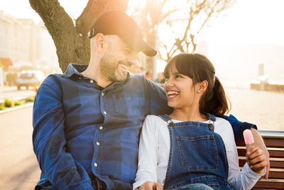 Cheerful father and daughter sitting on bench in city