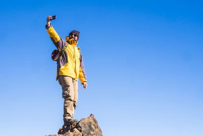 Low angle view of man taking selfie while standing on rock against sky