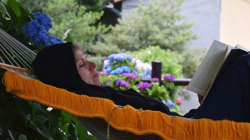 Side view of woman reading book while lying in hammock