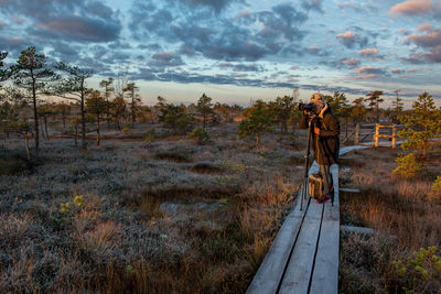 Woman photographing while standing on footbridge against sky