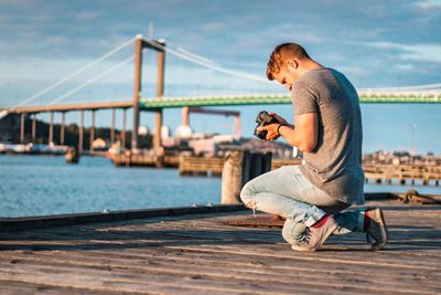 Man holding camera on pier by bridge over sea