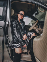 A girl wearing a polkadot dress, leather jacket, sun glasses and high boots sitting in a car 