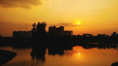 Silhouette buildings by lake against sky during sunset