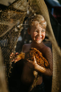 Smiling shirtless boy holding chicken while standing outdoors