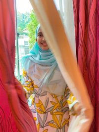 Young woman wearing hijab while peeking through curtain