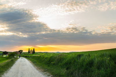 Dirt road amidst field against sky during sunset