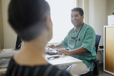 Happy male doctor looking at patient while using computer in clinic