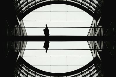 Symmetrical image of silhouette person walking on footbridge amidst buildings against clear sky