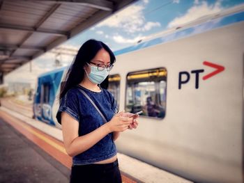 Side view of asian woman in eyeglasses and surgical face mask using smartphone at railway platform.