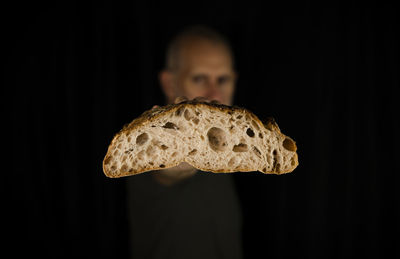 Close-up portrait of adult man holding bread cut in half against black background