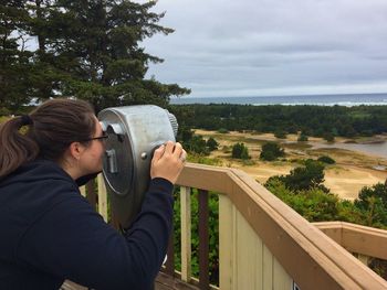 Side view of young woman looking through coin-operated binoculars against sky