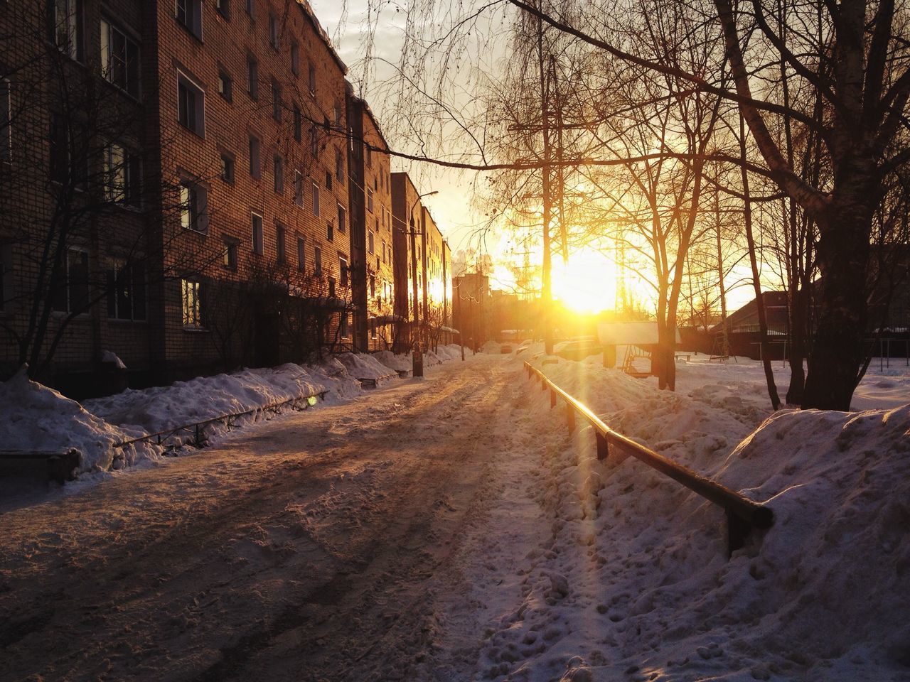 sunset, snow, sun, winter, cold temperature, the way forward, built structure, sunlight, season, bare tree, orange color, sunbeam, architecture, covering, nature, sky, building exterior, lens flare, tranquility, transportation