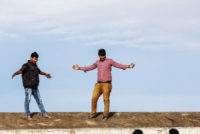 Men with arms outstretched standing on retaining wall against sky