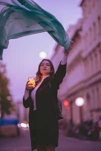 Portrait of beautiful woman moving scarf while holding illuminated jar in city at dusk
