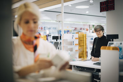 Senior female customer standing at checkout looking at owner in pharmacy store