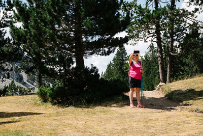 Hiker woman wearing straw hat, shorts and backpack on the path in a forest of pine trees stands up taking a photo of the nature while enjoys the natural environment around. horizontal photo.