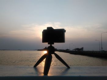 Digital camera with tripod on railing by sea against sky during sunset